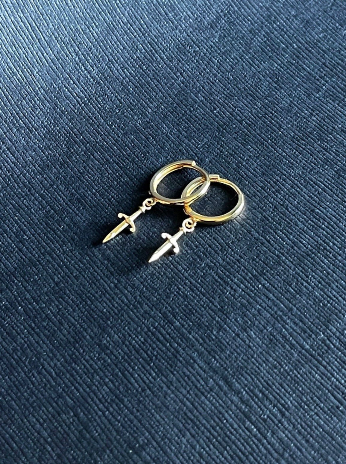 14K Gold small smooth Huggie Hoop Earrings with small Dangle Dagger Charm on a black textured background by  LeSilveStone
