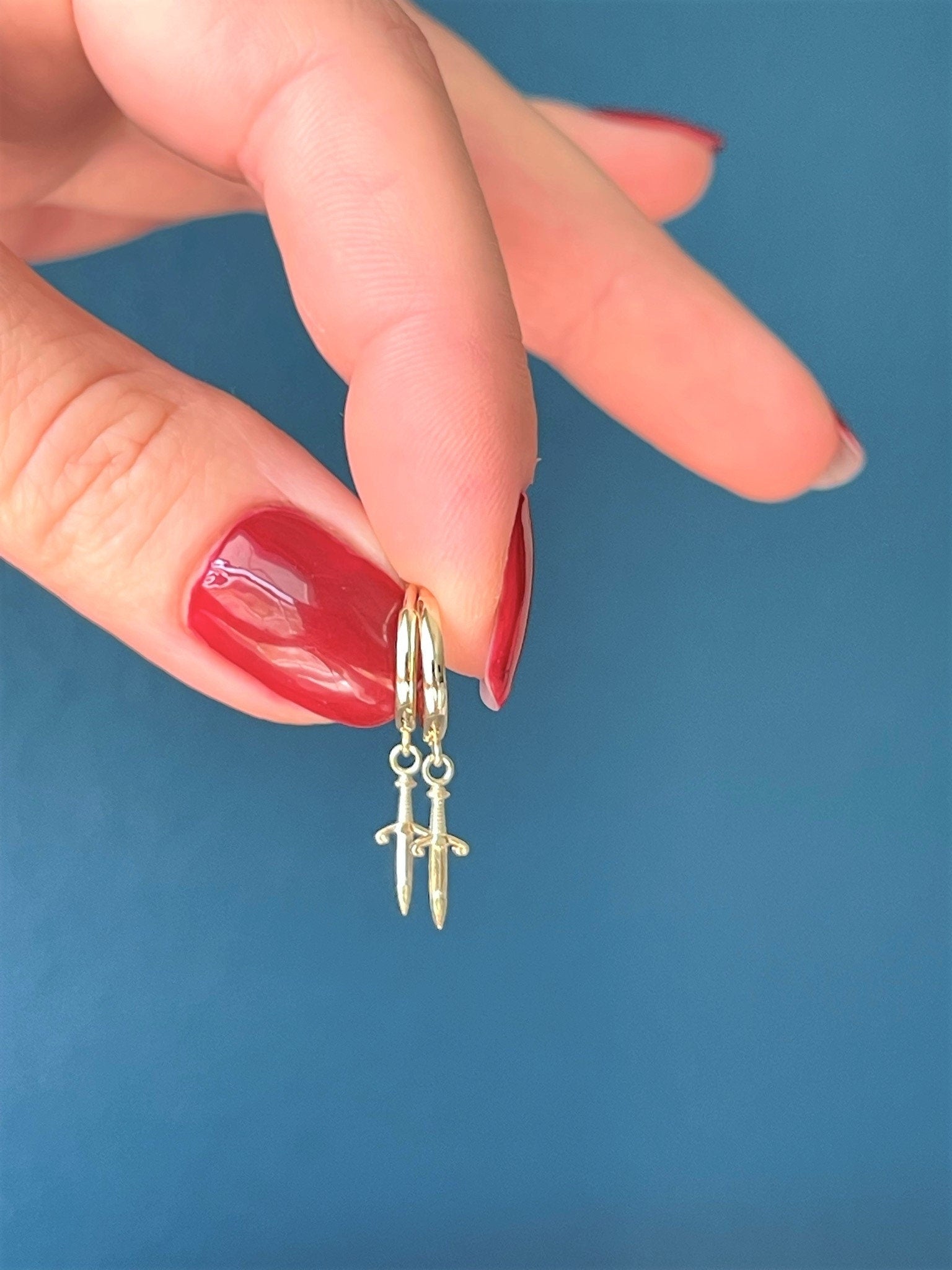 14K Gold Huggie Earrings with Dangle Dagger Charm held by a woman's fingers on a blue background by LeSilveStone