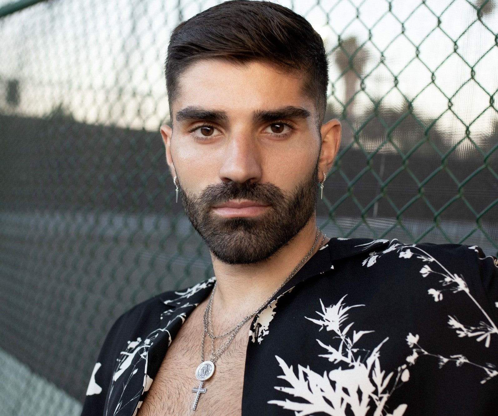 Unisex Womens and Mens Jewelry classic take on streetstyle Handcrafted in the USA dangle earrings huggie hoops and chain necklaces modeled on a male model in open black and white shirt  with a chain link fence in the background